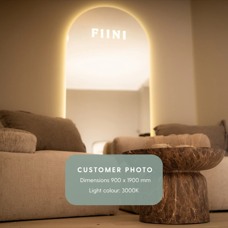 An arched backlit mirror on a beige wall with a text logo etched on to the upper end. The logo says "Fiini"