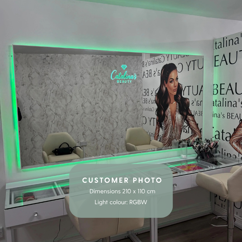 A square frameless mirror with RGBW backlights. Mirror is mounted on a wall in front of a beauty desk. The mirror has a text logo on the upper edge reading "Catalina's beauty".